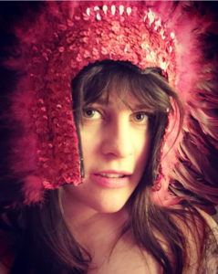 Face of a white, femme-ish person with dark brown hair (and some silver!) with bangs that extend over their thin, high-arching, dark brown eyebrows, and extends over their naked shoulders and onto their chest. Green eyes, open-mouthed pink slight smile. They are wearing a Vegas-style headdress made of sequins and feathers in various shades of pink, purple, and red.